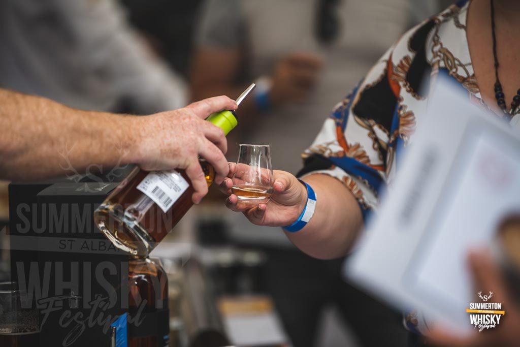 An exhibitor pouring a measure of whisky into a tasting glass held by a visitor at a Summerton whisky festival.